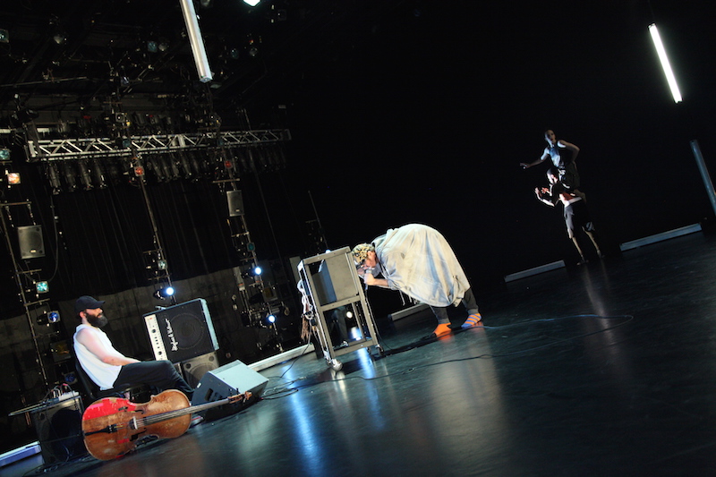 Musician Chris Lancaster projecting into the screen of an old TV. His cello lays on its side next two a sitting Nick Bruder.
